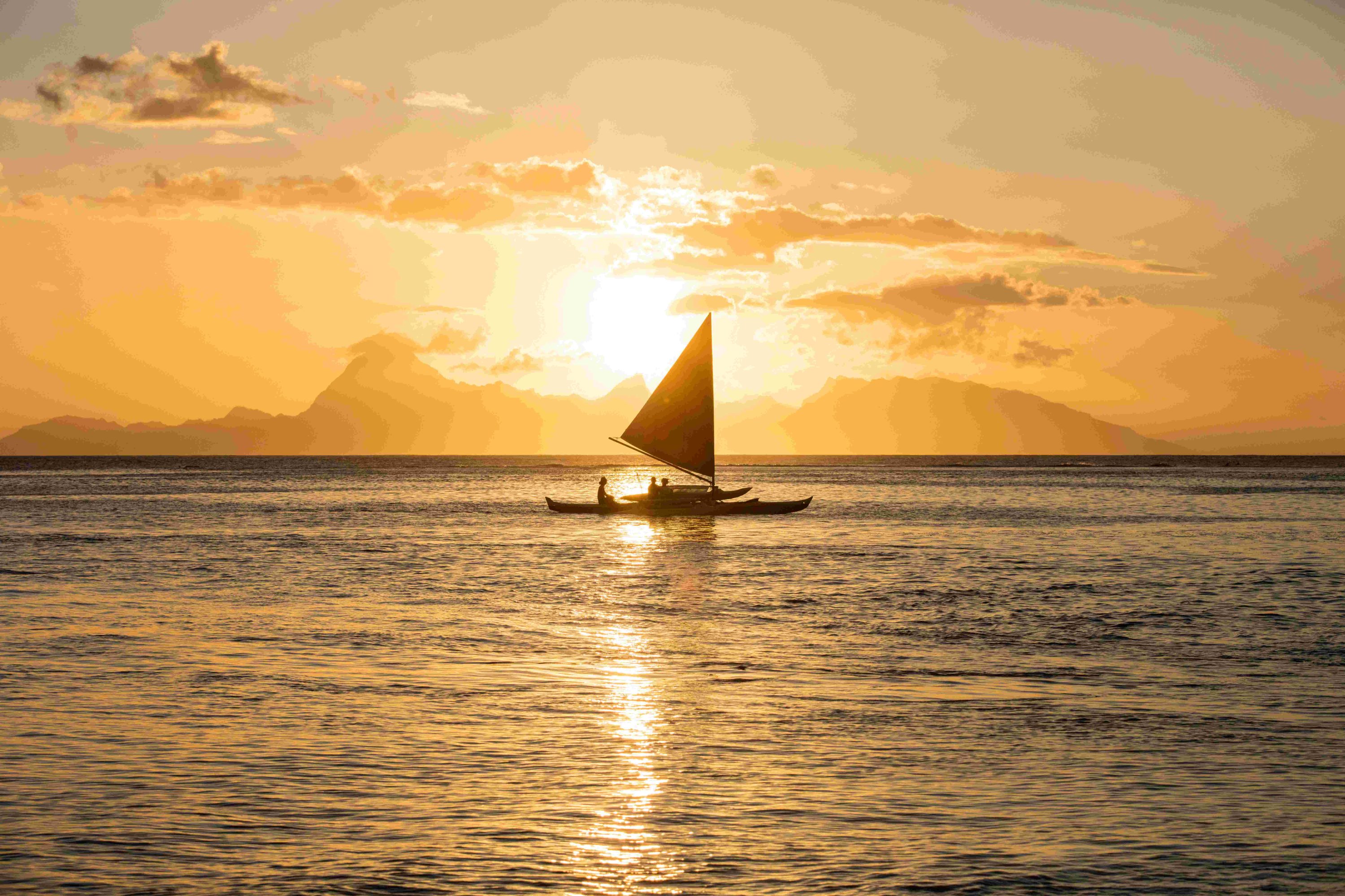 Pirogue à voile traditionnelle qui Navigue sur le lagon de Tahiti au coucher du soleil, avec derrière elle l'île de Moorea.